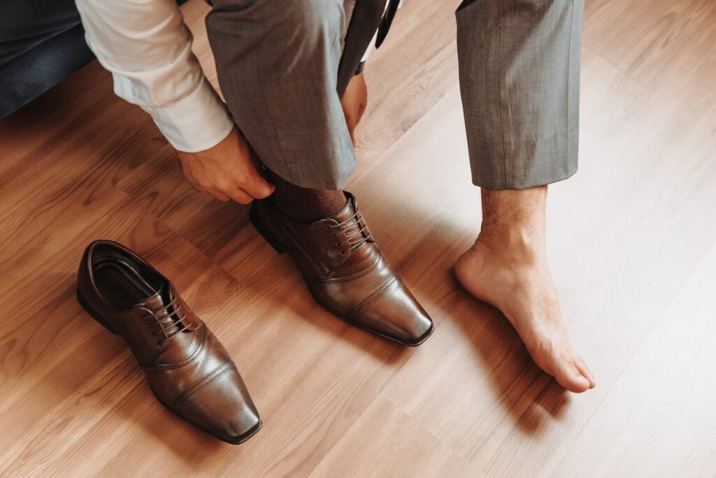 Close-up of a man in a suit putting on brown leather dress shoes indoors on a wooden floor.
